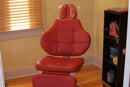Red dental chair in orthodontic office with wooden flooring and shelving unit.
