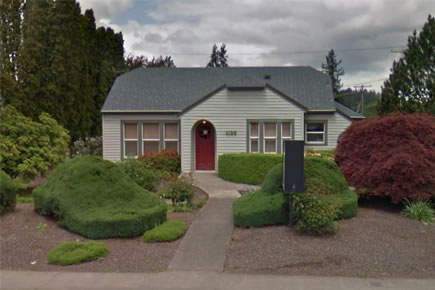 Small gray house with red door and landscaped front yard surrounded by evergreen trees.