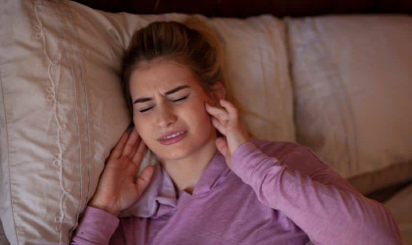 Woman in purple shirt lying on bed, touching ear, appearing uncomfortable or in pain.