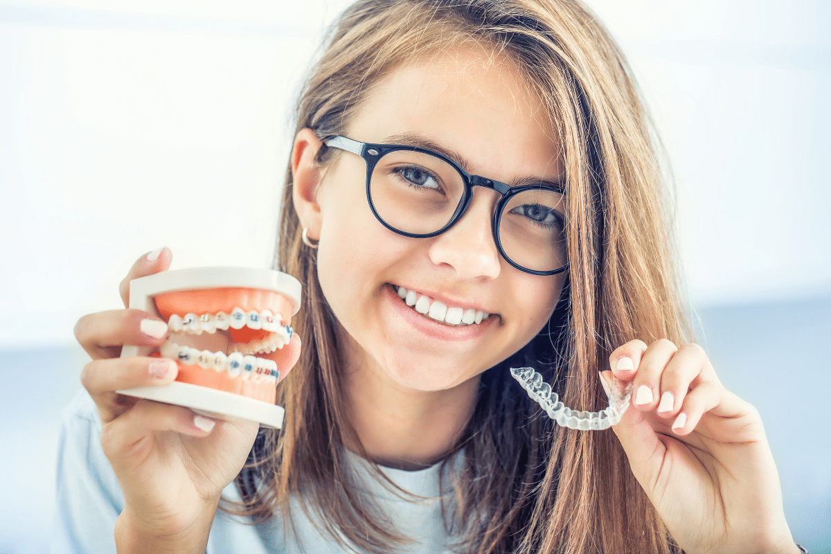 Young girl wearing glasses smiling while holding dental model and clear aligners indoors.